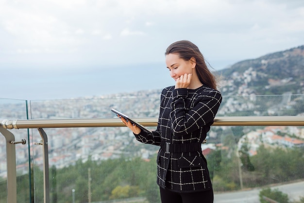 Blogger girl is looking at planshet computer in her hand by holding hand on cheek against the background of city view