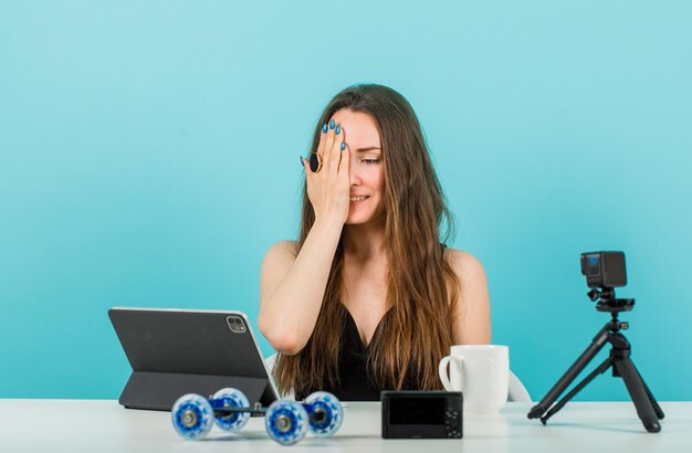 Blogger girl is looking at tablet by covering half of face with hand on blue background