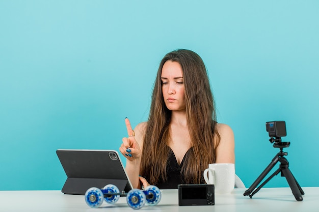 Blogger girl is looking at tablet camera by showing a minute gesture on blue background