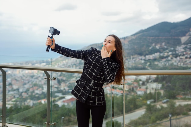 Blogger girl is showing kiss gesture by holding hand on lips to camera against the background of city view