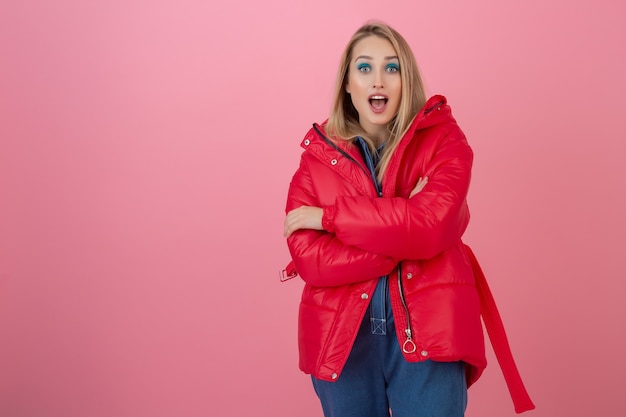 Free photo blond attractive active woman posing on pink wall in colorful winter down jacket of red color, having fun, warm coat fashion trend, surprised shocked face expression