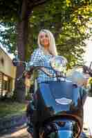 Free photo blond female in casual clothing possing on moto scooter over green trees background.