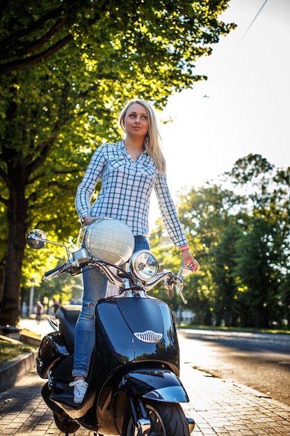 Free Photo blond female in casual clothing possing on moto scooter over green trees background.