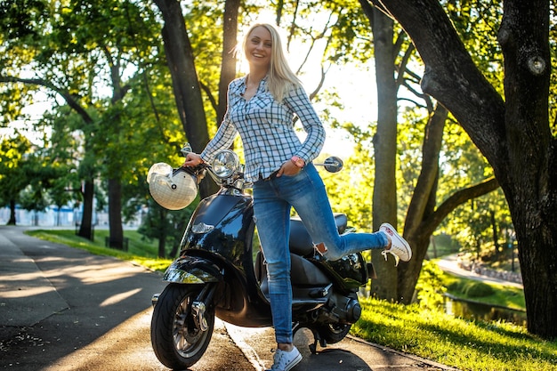 Free photo blond female in casual clothing possing on moto scooter over green trees background.