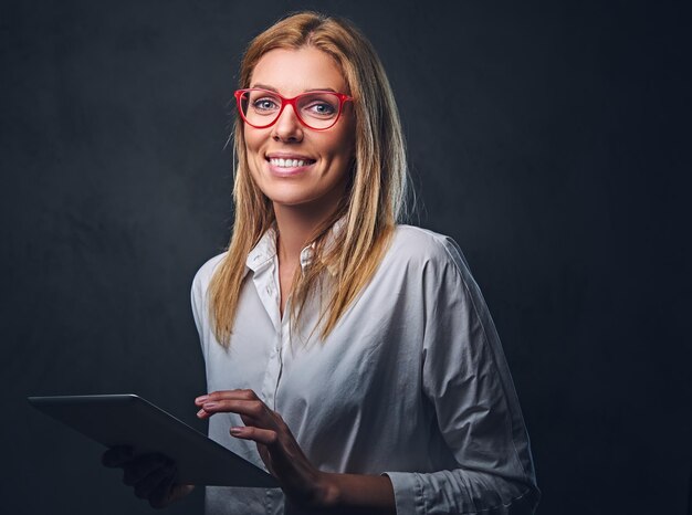 A blond female dressed in a white shirt and eyeglasses holds tablet PC on grey background.