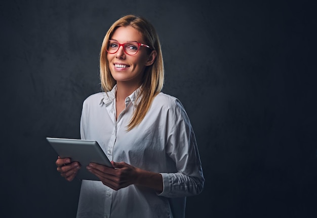 A blond female dressed in a white shirt and eyeglasses holds tablet PC on grey background.