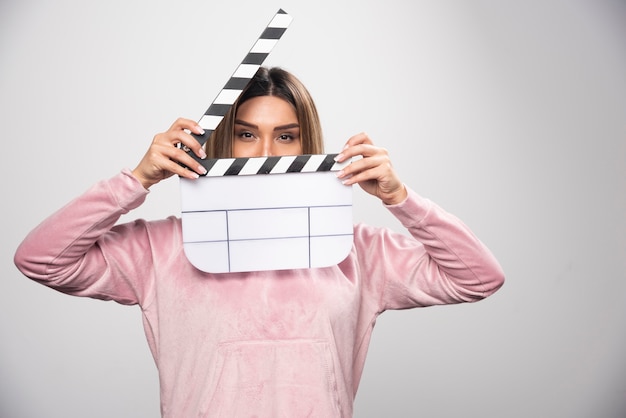 Free Photo blond lady in pink sweatshier holding a blank clapper board and looking through it.