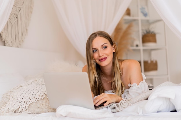 Blond smiling woman  lying on cozy bed  and using laptop for online shopping.