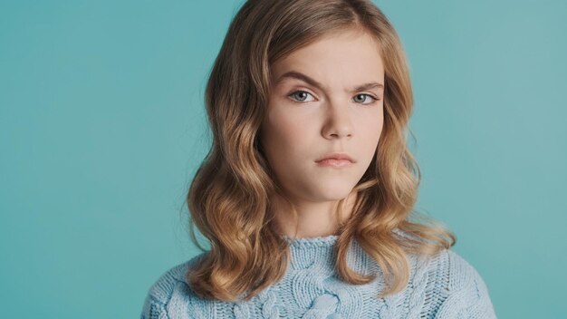 Blond teenage girl raising eyebrow on camera looking serious over blue background. Suspicion expression