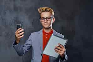 Free photo blond teenager boy in eyeglasses holds tablet pc and smartphone.