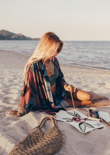 Free photo blond woman drawing watercolor flower by brush on the beach