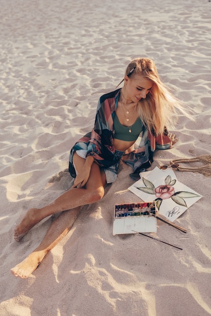 Free photo blond woman drawing watercolor flower by brush on the beach