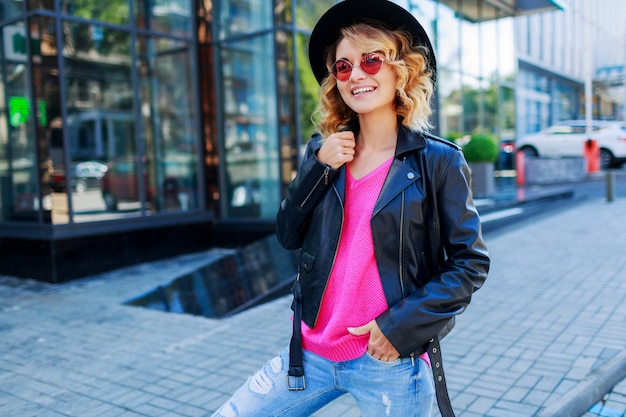 Free photo blond woman posing on modern streets. stylish autumn outfit, leather jacket and knitted sweater. pink sunglasses.