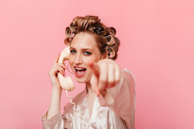 Blond woman in silk home outfit is talking on phone and points her finger at front against isolated wall