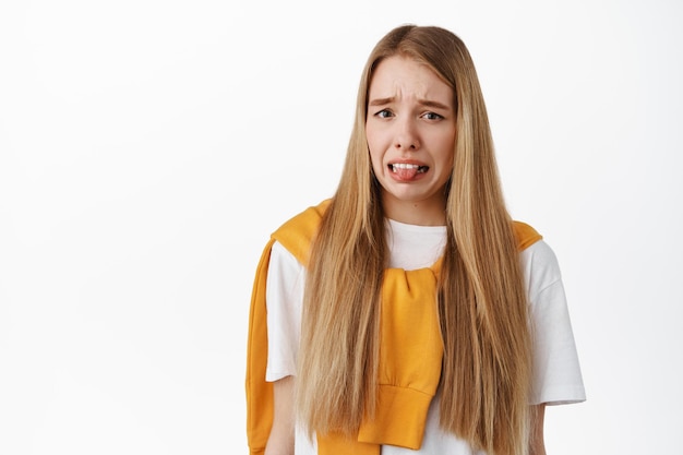 Free photo blond young woman feeling disgusted, showing tongue and grimacing from dislike, staring at something gross disgusting, standing against white background