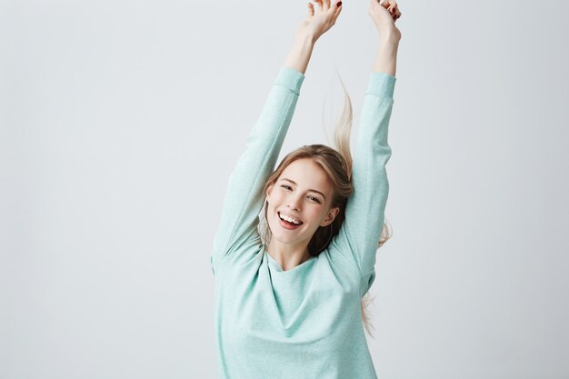 Blonde beautiful young woman in light blue top stretching arms up in cheerful mood like celebrating victory. Broadly smiling female showing white teeth and positive emotions, having fun indoors.