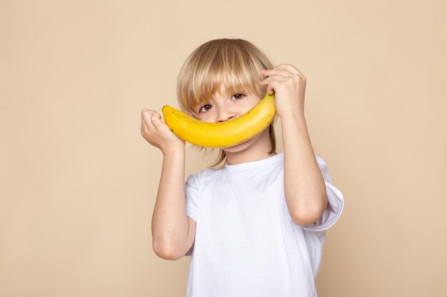 blonde boy smiling cute sweet with banana in white t-shirt on pink desk