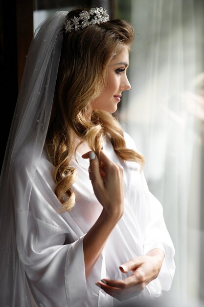 Blonde bride in the crown stands before bright window in the morning 