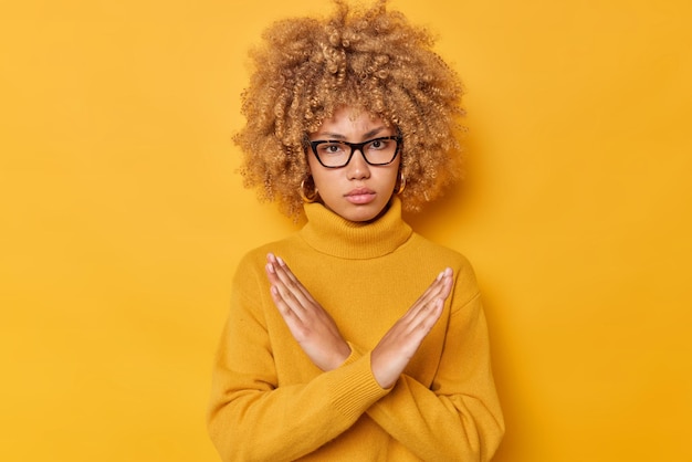 Blonde curly haired woman shows cross makes stop gesture says no with angry expression declines something wears spectacles and long sleeved jumper isolated over yellow background. Its taboo.