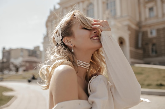 Free Photo blonde curly young woman smiles sincerely outdoors happy charming girl in white blouse and pearl necklace poses in city center