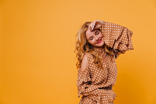 Blonde female model posing on yellow wall with relaxed smile. Adorable young woman with red lips chilling