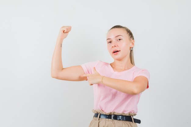 Blonde female pointing at muscles of arm in t-shirt, pants and looking confident. front view.