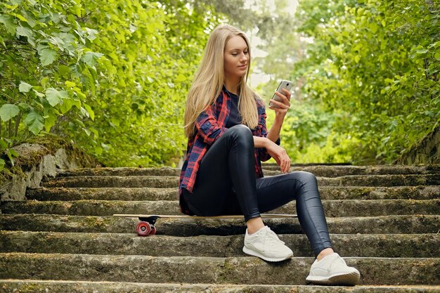 Blonde female using a smartphone and sits on Longboard on the steps.