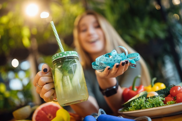 Free photo blonde fithess girl sit on the table and holding fresh smoothie and measure, diet time concept.