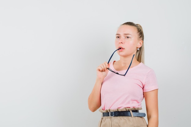 Blonde girl biting glasses in t-shirt, pants and looking thoughtful , front view.