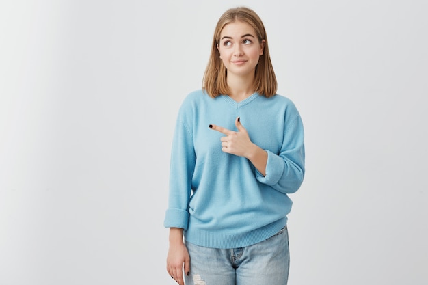 Blonde girl in blue sweater looking with her dark eyes aside pointing with index finger at copy space advertising something. Woman posing against gray wall with copy space for text or promotion