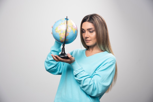 Free Photo blonde girl in blue sweatshirt holding a globe and checking the earth map attentively.