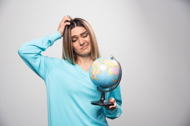 Free photo blonde girl in blue sweatshirt holds a globe and looks uncertain and confused.