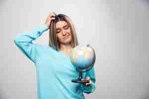 Free photo blonde girl in blue sweatshirt holds a globe and looks uncertain and confused