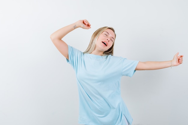 Blonde girl in blue t-shirt stretching and yawning, keeping eyes closed and looking sleepy , front view.