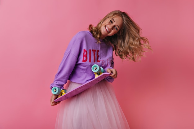 Free Photo blonde girl in casual shirt holding skateboard and laughing. charming female model expressing sincere emotions on pink.