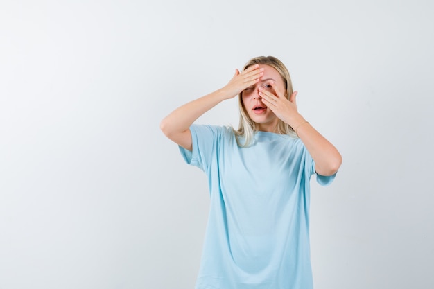 Blonde girl covering eyes with hands, looking through fingers in blue t-shirt and looking pretty , front view.