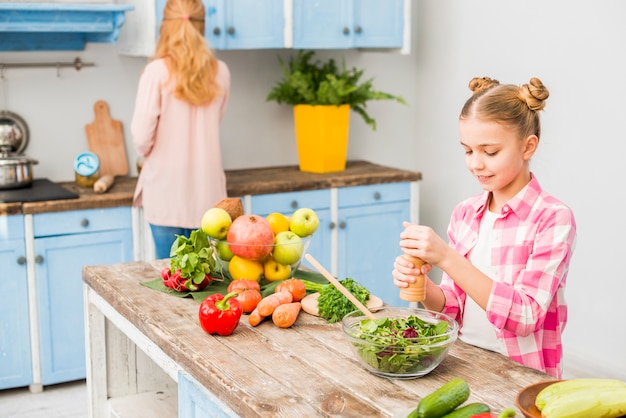 Blonde girl grinding the pepper in salad bowl with her mother in the kitchen