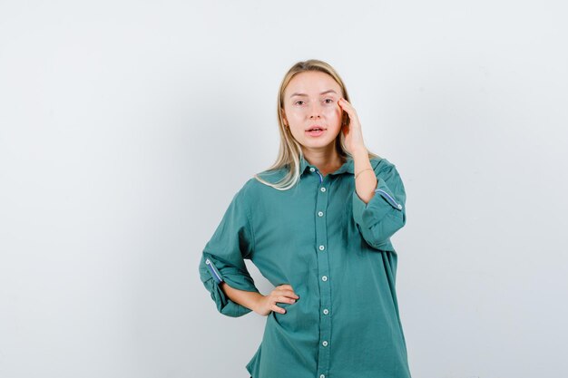 Blonde girl holding hand on waist while putting hand on cheek in green blouse and looking serious.