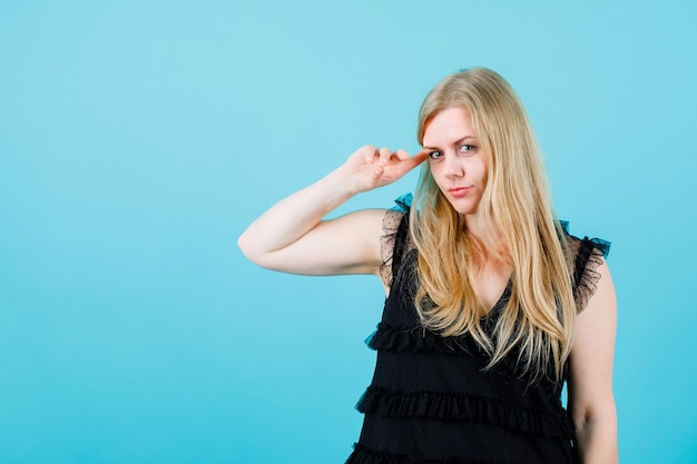 Blonde girl is looking at camera by holding forefinger on temple on blue background