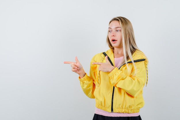 Blonde girl in pink t-shirt and yellow jacket pointing left with index fingers and looking alluring