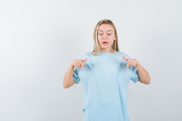 Free Photo blonde girl pointing down with index fingers in blue t-shirt and looking focused. front view.