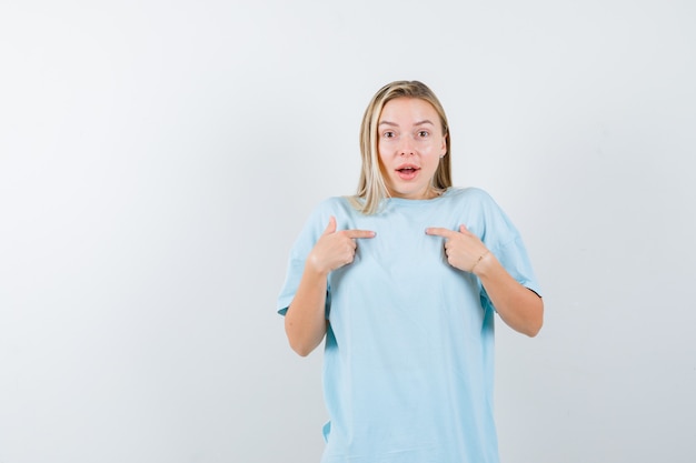 Blonde girl pointing at herself with index fingers in blue t-shirt and looking surprised. front view.