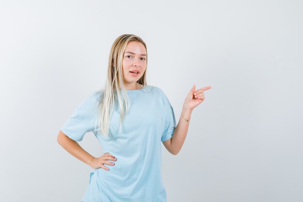 Blonde girl pointing right with index finger, holding hand on waist in blue t-shirt and looking pretty , front view.