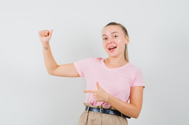 Blonde girl pointing at something pretended to be held in t-shirt, pants and looking happy , front view.