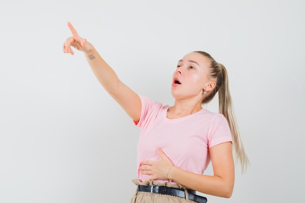 Blonde girl pointing up in t-shirt, pants and looking surprised , front view.