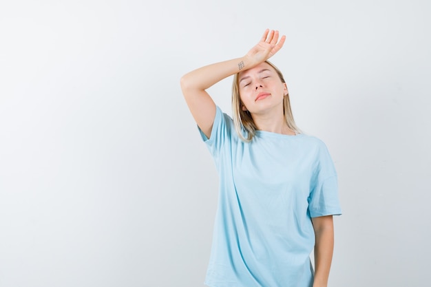 Blonde girl putting hand on forehead, having headache in blue t-shirt and looking exhausted. front view.