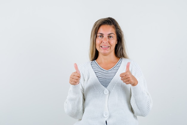 Free photo blonde girl showing double thumbs up in striped t-shirt, white cardigan and jean pants and looking happy. front view.