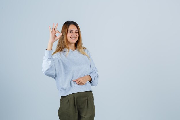 Blonde girl showing ok sign in olive green blue sweatshirt and pants and looking radiant , front view.