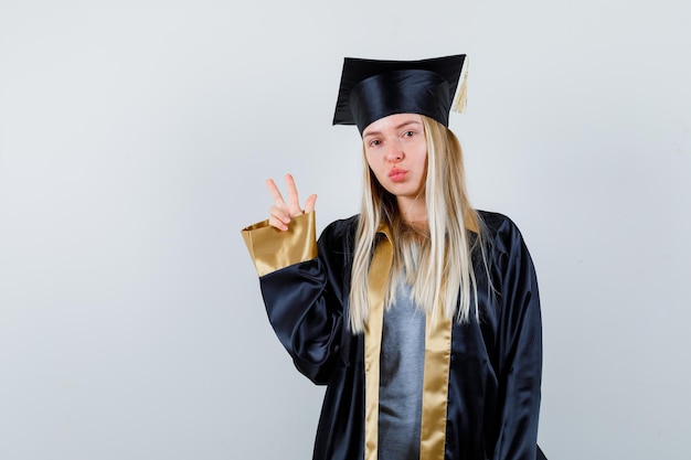 Free photo blonde girl showing peace gesture in graduation gown and cap and looking cute