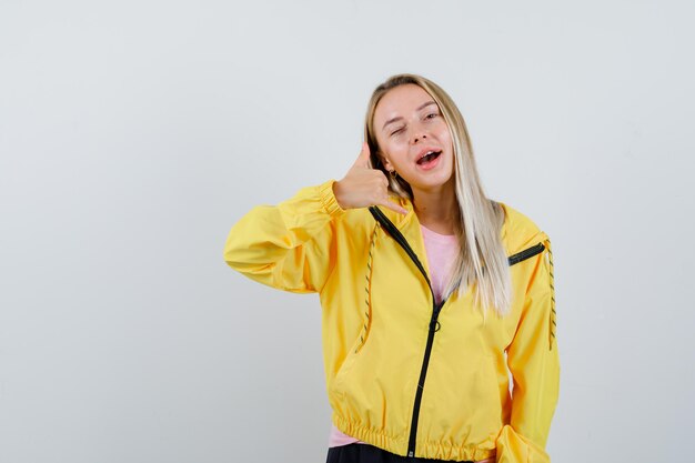 Blonde girl showing phone gesture in yellow jacket and looking confident.